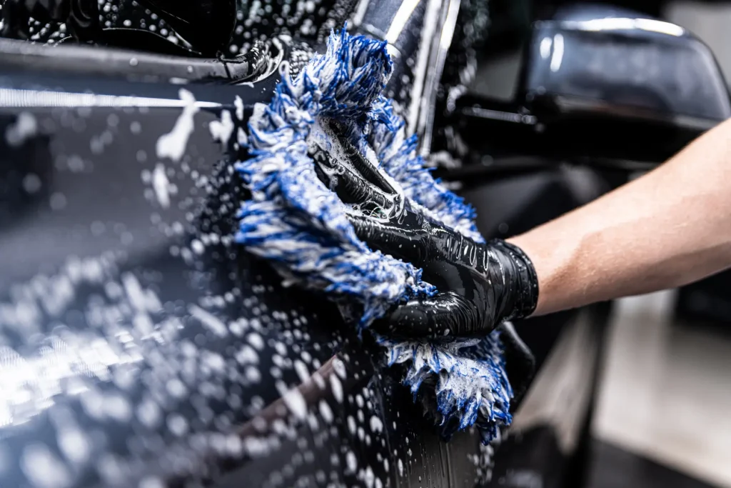 Eine Person mit schwarzen Handschuhen reinigt ein schwarzes Auto mit blauem Waschhandschuh